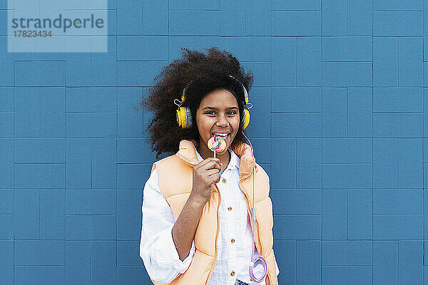 Mädchen leckt Lutscher und hört Musik über kabellose Kopfhörer vor blauer Wand
