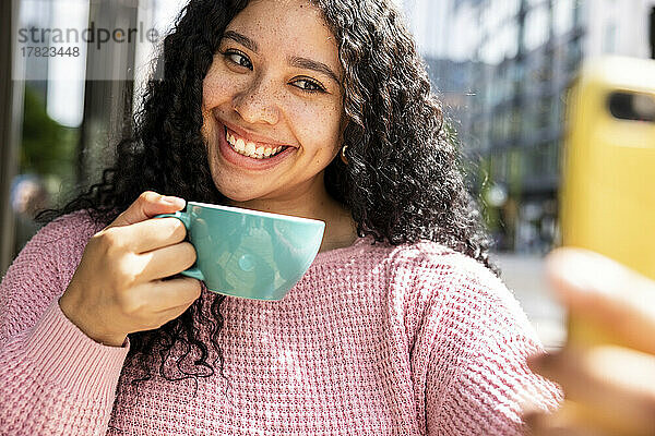 Glückliche Frau hält eine Kaffeetasse in der Hand und macht an einem sonnigen Tag ein Selfie mit dem Smartphone
