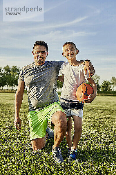 Glücklicher Mann kniet neben seinem Sohn und hält Rugbyball auf dem Sportplatz