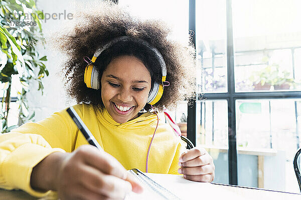 Fröhliches Mädchen mit kabellosen Kopfhörern macht Hausaufgaben im Café