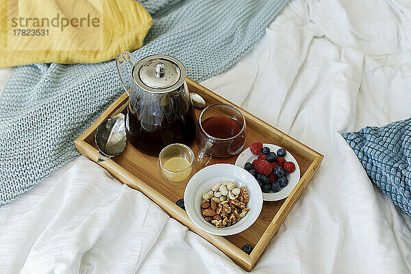 Teekanne in Holztablett mit Beeren und Nüssen auf dem Bett