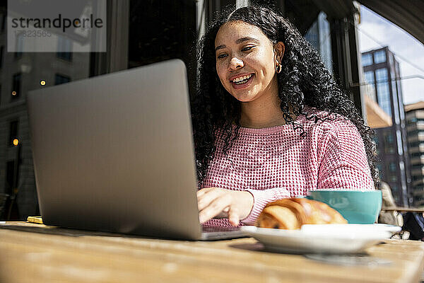 Glückliche Frau mit schwarzen Locken benutzt Laptop im Straßencafé