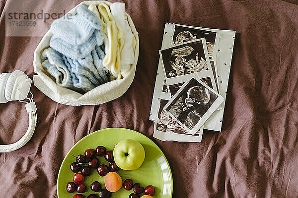 Sonogramme mit Babykleidung und frischem Obst auf dem Bett