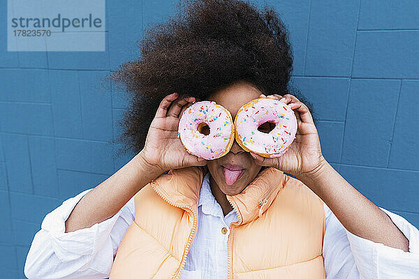 Mädchen streckt die Zunge heraus und bedeckt Augen mit Donuts vor blauer Wand