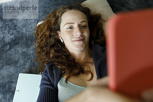 Smiling woman using phone lying on carpet at home