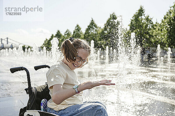 Fröhliches Mädchen mit Behinderung sitzt im Rollstuhl und spielt am Brunnen
