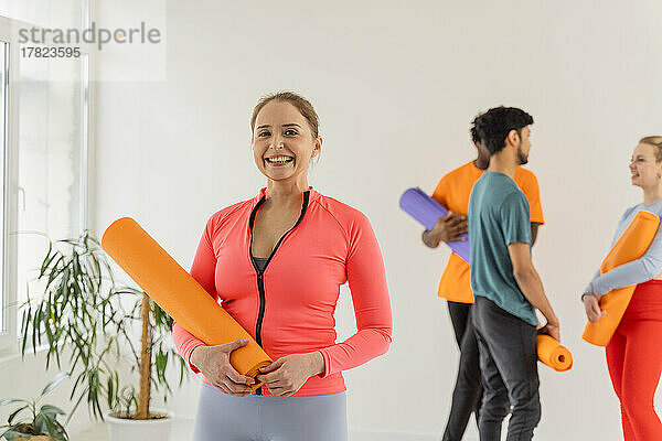 Lächelnde Frau hält Trainingsmatte mit Freunden und unterhält sich im Yoga-Studio