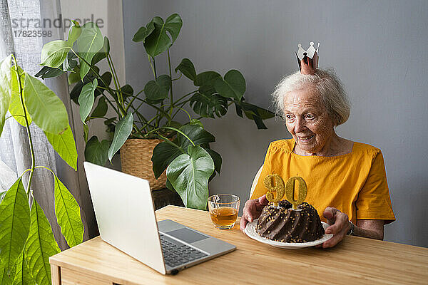 Glückliche Seniorin zeigt Geburtstagstorte bei Videoanruf über Laptop zu Hause
