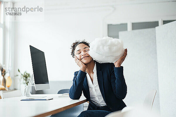 Lächelnde Geschäftsfrau mit geschlossenen Augen  die eine Wolke im Büro hält