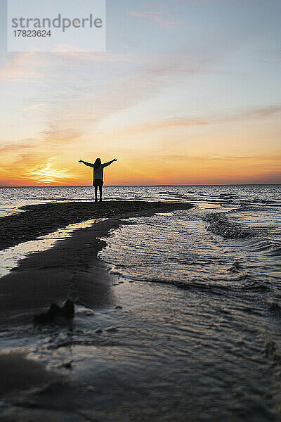Junge Frau mit ausgestreckten Armen steht bei Sonnenuntergang an der Küste