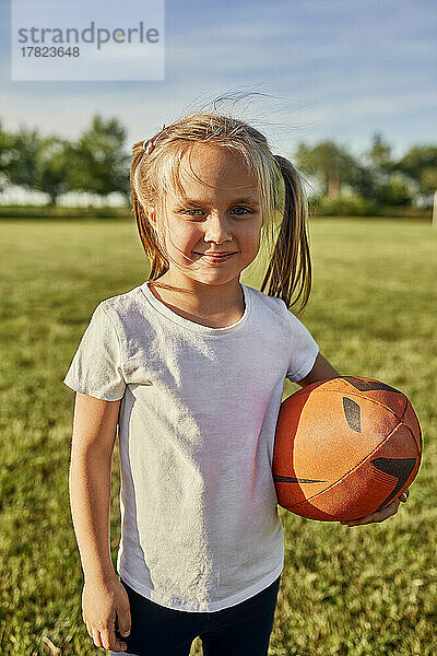 Lächelndes blondes Mädchen mit Rugbyball an einem sonnigen Tag