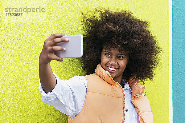 Glückliches Mädchen  das Selfie mit dem Smartphone vor der gelben Wand macht