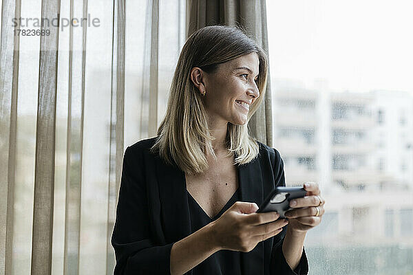 Lächelnde Geschäftsfrau hält Mobiltelefon und schaut durchs Fenster