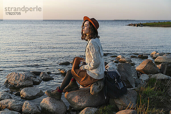 Junge Frau mit Hut sitzt auf einem Felsen an der Küste