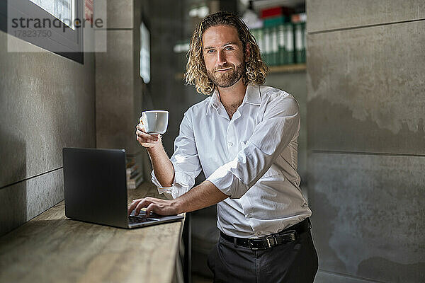 Führungskraft hält Kaffeetasse mit Laptop auf dem Tisch im Büro