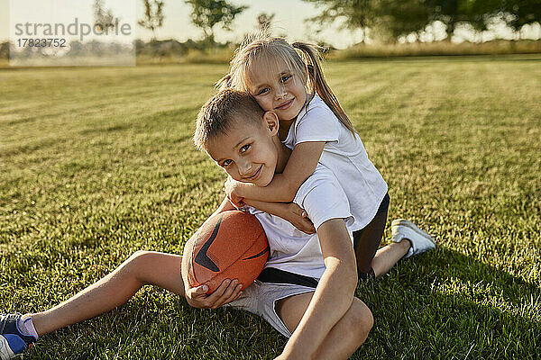 Lächelndes blondes Mädchen umarmt Bruder  der mit Rugbyball auf dem Sportplatz sitzt