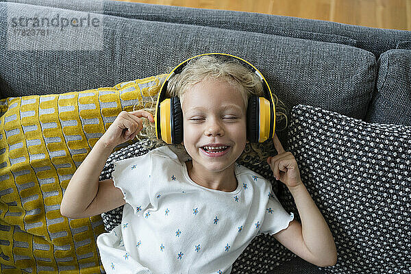 Glückliches Mädchen mit geschlossenen Augen  das zu Hause auf dem Sofa Musik über kabellose Kopfhörer hört