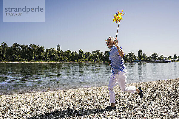 Glücklicher älterer Mann mit Windradspielzeug aus Papier  der am Flussufer läuft
