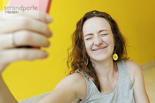 Glückliche Frau mit geschlossenen Augen macht ein Selfie mit dem Mobiltelefon vor einer gelben Wand