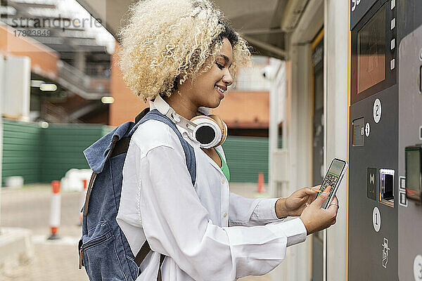 Lächelnde Frau scannt QR-Code per Smartphone am Ticketautomaten