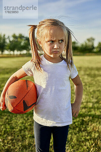 Unzufriedenes blondes Mädchen mit Rugbyball steht an einem sonnigen Tag auf dem Sportplatz