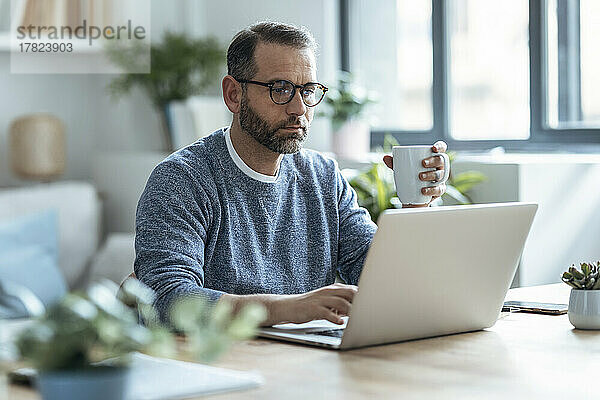 Geschäftsmann hält Kaffeetasse in der Hand und arbeitet zu Hause am Laptop