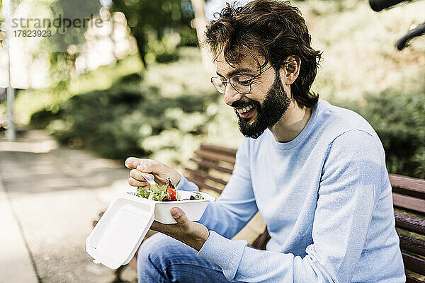 Lächelnder Mann mit Salat sitzt auf Bank im Park