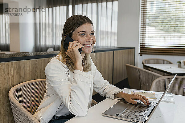 Lächelnde Geschäftsfrau  die mit einem Laptop im Restaurant sitzt und mit dem Handy telefoniert