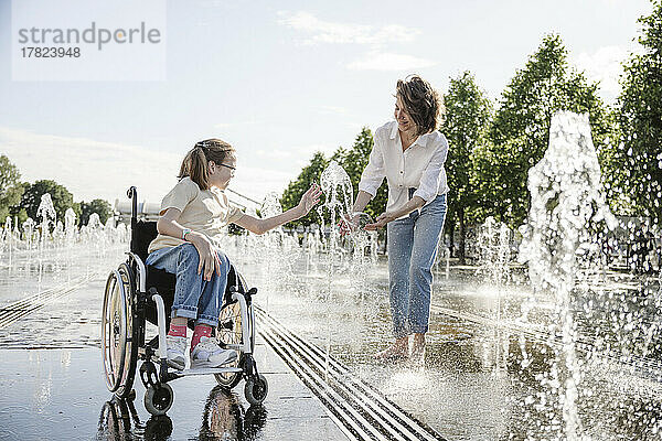 Mädchen im Rollstuhl spielt mit Mutter im Park am Brunnenwasser
