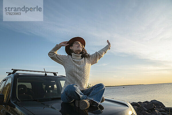 Lächelnde Frau mit Hut sitzt auf der Motorhaube eines Autos