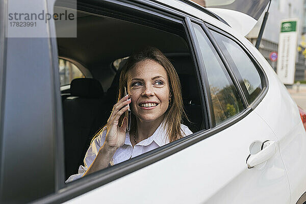 Lächelnde Geschäftsfrau  die am Telefon spricht und durch das Autofenster schaut