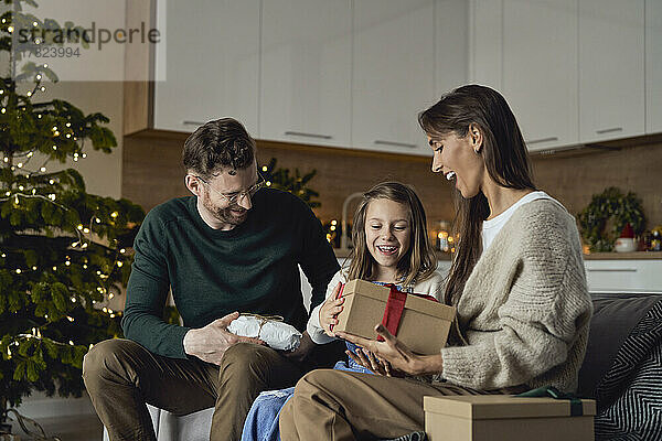 Glückliche Eltern geben ihrer Tochter  die auf dem Sofa sitzt  ein Weihnachtsgeschenk