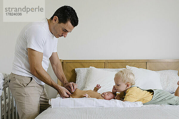 Boy looking at man buttoning baby's rompers on bed at home