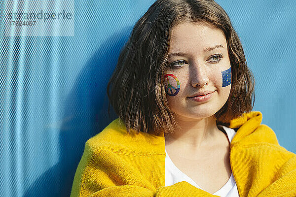 Nachdenkliches Teenager-Mädchen mit Friedenssymbol und EU-Farbe auf den Wangen vor blauer Wand