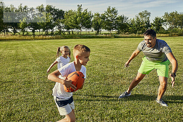 Vater spielt an einem sonnigen Tag mit Sohn und Tochter Rugby auf dem Sportplatz