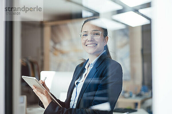 Glückliche Geschäftsfrau mit Tablet-PC durch Glas Büro gesehen