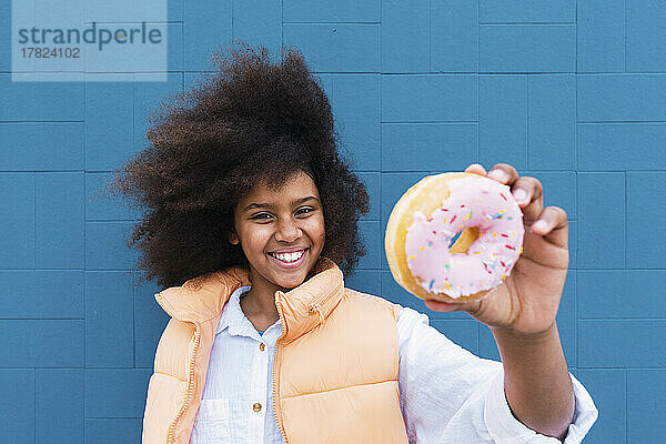 Smiling girl showing doughnut standing in front of blue wall