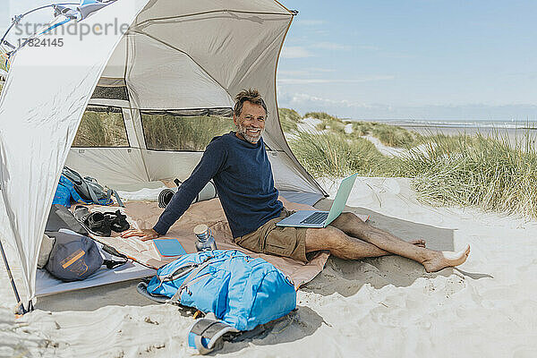 Glücklicher Freiberufler mit Laptop  der an einem sonnigen Tag im Zelt am Strand sitzt