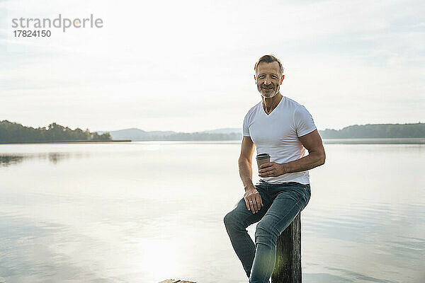Glücklicher reifer Mann mit Einwegkaffeetasse  der auf einem Pfosten vor dem See sitzt