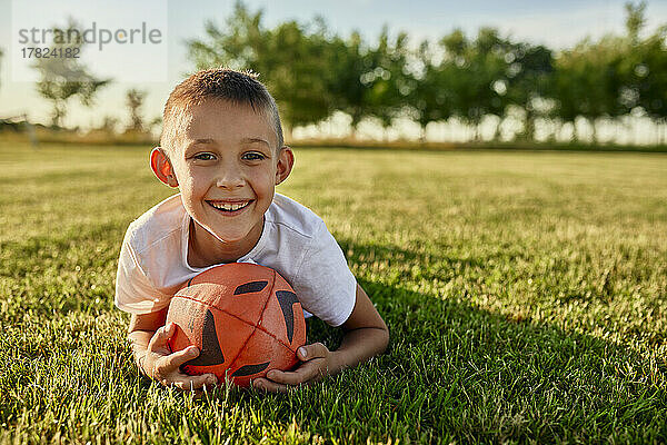 Glücklicher Junge mit Rugbyball  der an einem sonnigen Tag auf dem Sportplatz liegt