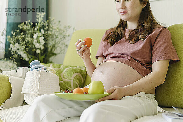 Schwangere Frau isst gesunde Früchte im Wohnzimmer