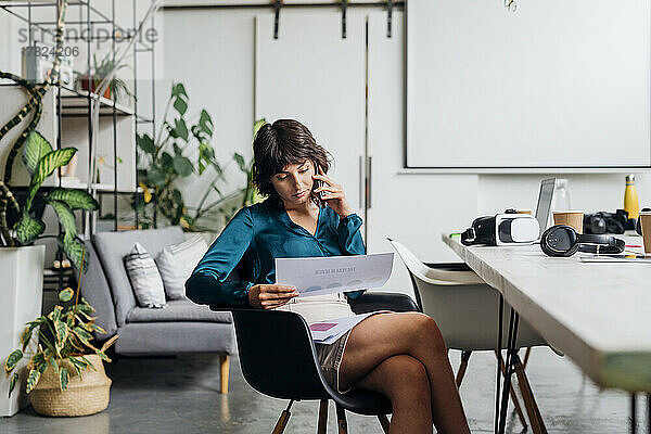 Junge Geschäftsfrau mit Dokumenten  die im Büro auf dem Smartphone spricht