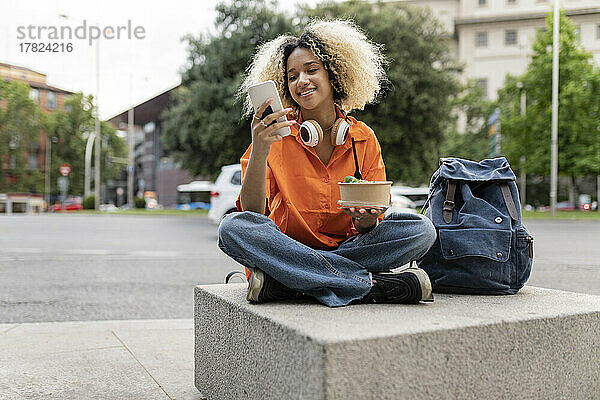 Lächelnde Frau mit Smartphone und Brotdose in der Hand  die mit gekreuzten Beinen auf dem Sitz sitzt