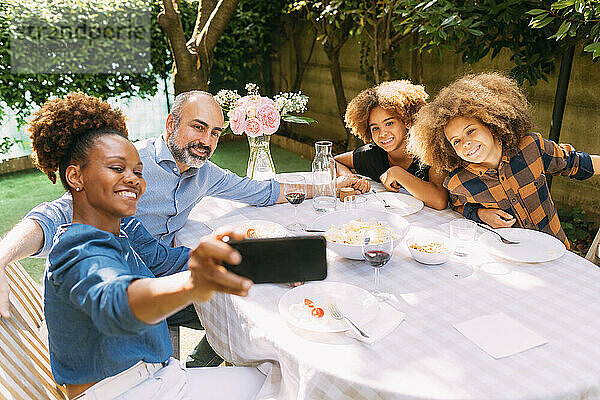 Lächelnde Frau macht ein Selfie mit der Familie per Smartphone am Esstisch im Hinterhof