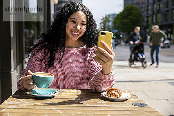 Glückliche Frau hält eine Kaffeetasse in der Hand und führt einen Videoanruf über ihr Mobiltelefon im Straßencafé