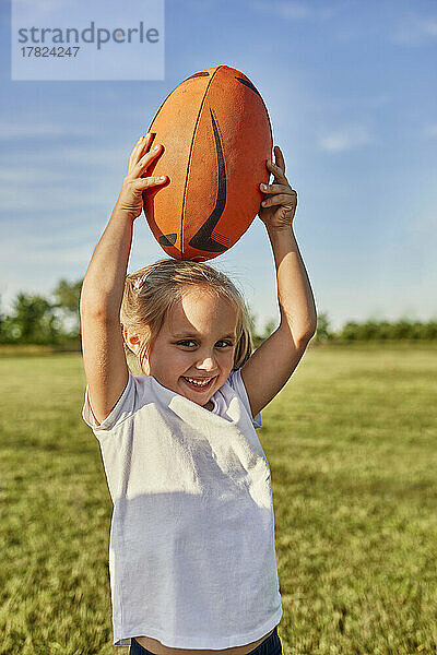 Glückliches blondes Mädchen trägt Rugbyball auf dem Kopf auf dem Sportplatz
