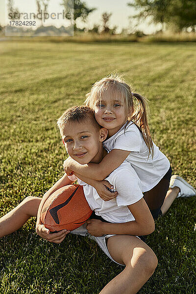Lächelndes Mädchen umarmt Bruder  der an einem sonnigen Tag mit Rugbyball auf dem Sportplatz sitzt