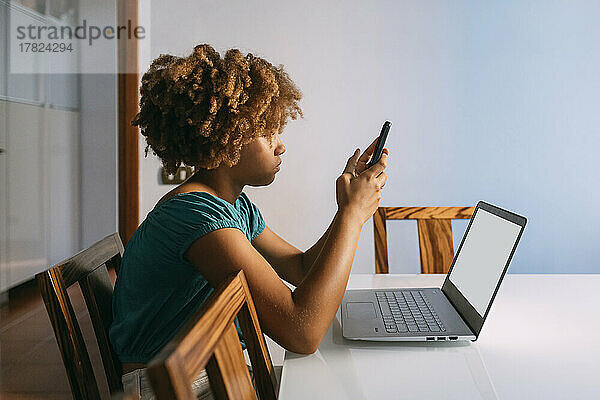 Mädchen sitzt mit Laptop und nutzt Smartphone zu Hause