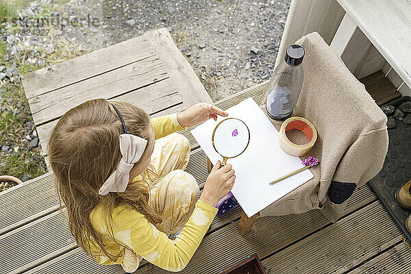 Girl observing flower petal though magnifying glass sitting at porch