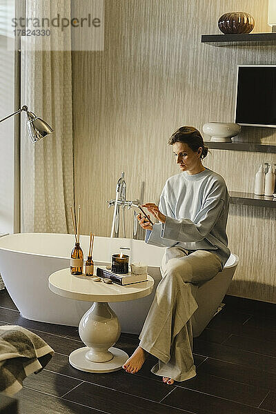 Frau sitzt auf Badewanne im Badezimmer und benutzt Smartphone